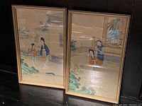 Pair of framed Chinese paintings on silk leaned against a wall on a floor, showing traditional figures and landscape details.