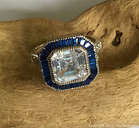 Front view of sterling silver ring with large clear square-cut central stone surrounded by blue lab created sapphire baguette stones on a wooden background.