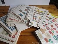 Photo of loose pages of stamp collections arranged in multiple piles on table, showing many stamps affixed on pages with labels.