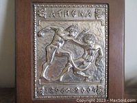 Frontal view of the silver plaque on wooden frame showing detailed athlete relief, text 'ATHENA' and '1896-2004' with decorative elements.