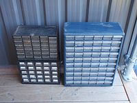 Three plastic hardware organizers of different sizes and colors with multiple small drawers on wooden floor against metal wall.