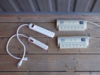 Four power bars on wooden floor showing top and cords. Two white 6-outlet strips and two beige 8-outlet strips with switches.