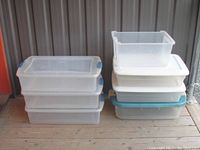 Lot of seven stacked clear plastic storage bins with lids, showing various sizes and colors including clear lids with blue latches, white lids, and a blue lid, plus a small bin without a lid on top in one stack