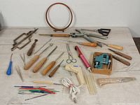 Wide shot of multiple leather working tools arranged on a flat surface including glue gun, punches, scissors, embroidery hoop, and pliers.