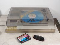 Front perspective of Sharp SG-170X turntable stereo radio cassette player with dust cover down, showing blue Sharp branded platter mat and surrounding controls with two blank cassette tapes and black LP cleaner brush in front.