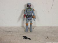 Front view of vintage Boba Fett figure standing on surface with original black blaster in front, showing paint wear and detailing on figure