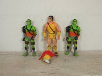 Three action figures displayed side by side: two green orc warriors and one gladiator warrior without helmet in front of detached gladiator helmet with red plume.
