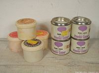 Nine containers of powder paint: four tall metal tins labeled Winsor & Newton New Art Powder Colour and five smaller plastic jars labeled Mona Lisa powder paint in different colors.