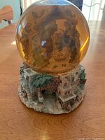 Photo showing the front and side view of the musical snow globe with nativity scene inside a golden-tinted water globe, mounted on a stone and palm tree textured base.