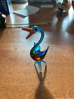 Front-left view of the blue glass bird figurine showing orange beak and body with blue legs and head, placed on wooden surface.