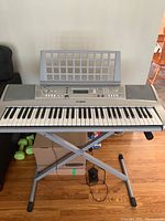 Front view of Yamaha PSR-E303 keyboard on adjustable X-style stand with music rest, foot pedal and power adapter visible underneath