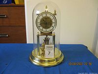 Full view of gold-tone anniversary clock under glass dome on blue background with MaxSold lot card.