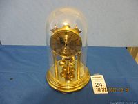 Front view of brass anniversary clock under glass dome, showing dial and rotating pendulum.