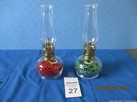 Two glass oil lamps side by side on a blue fabric surface, one with red beads and one with green beads inside the base, with clear chimneys and brass burners visible.
