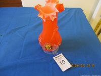 Full view of the orange Murano vase with ruffled top and decorative glass flowers around lower part, displayed on blue cloth.