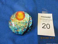 Top view of round glass paperweight featuring a bright orange sun and stylized tree design on blue background.