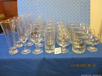 Full set of 28 Calgary Olympic glasses arranged on a blue cloth surface with a diamond plate background, showing multiple glass styles with gold rims and Olympic designs.