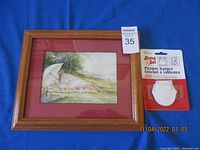 Framed picture with image of woman at picnic near water, showing frame and attached hangers.