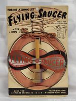 Front view of the 1950s Atomic Jet Flying Saucer toy mounted on original card backing, showing colorful retro graphics and toy design including a plastic flying saucer and wooden spool.