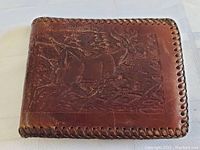 Front top view of brown leather wallet with detailed deer and forest tooled design and braided edge stitching.