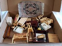 Vintage dollhouse furniture collection in cardboard box including a patterned rug, various wooden chairs, tables, cabinets and small furniture pieces with detail.