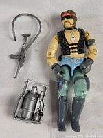 G.I. Joe action figure lying down with accessories including rifle, backpack oxygen tank, and grappling hook with rope around it.