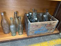 Glass bottles displayed inside and outside of wooden crate showing various bottle styles, closures, and embossed logos.