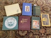 Photo of six vintage children's books varying in size and color, arranged on a patterned carpet. Titles include Slovenly Peter, The Little House, Orchids of New England, The Land of Pretend, When We Say: Hail Mary, and The Great Rain Robbery.