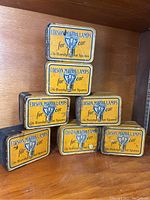 Seven antique rectangular tins stacked in a pyramid formation on wooden shelf. Each tin has a yellow label with blue and black text and an automotive light bulb illustration. Paint marks visible on sides and edges worn.