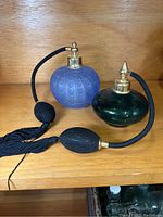 Both glass perfume bottles side by side on wooden shelf. One blue with swirl design, one green with flat shape. Each has gold-tone atomizer, black tubes and tassels.