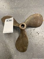 Top view of a bronze boat propeller lying on a concrete floor, showing three curved blades and central hexagonal hole with crack