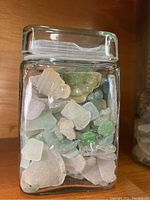 Front view of clear glass jar showing frosted sea glass pieces in various white, green, and light blue colors with some yellowish pieces.