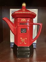 Front angle of red ceramic Victorian postbox shaped teapot with gold detailing and black base. Shows the spout and handle and royal crest design.