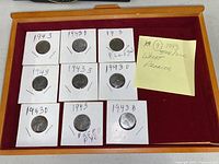 All nine 1943 steel/zinc wheat pennies laid out in a wooden display box inside individual cardboard holders with handwritten date and mint mark annotations.