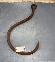 Large rounded iron hook with eyelet at one end and curved pointed hook at the other, laid on concrete floor showing surface rust and wear.