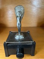 Front view of table lighter showing winged female figure on top and black rectangular base with ignition knob.