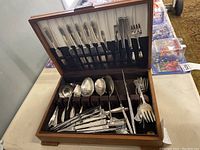 Open wooden box of silverplated flatware showing knives, forks, spoons in compartments and slots.