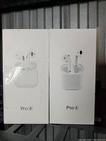 Photo shows two unopened white rectangular boxes of Pro 4 wireless earbuds placed side by side on a metal surface. Each box features an image of the earbuds and charging case on the front, with the label 'Pro 4' beneath the image.