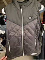 Photo shows black men's Medium heated vest with front zipper and two zippered pockets. Quilted bottom front panel and smooth upper chest area. Logo patch visible on chest.
