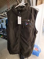 Black fleece women's heated vest hanging on a white plastic hanger. Visible tags and embroidered brand logos. Zippered pockets present.