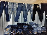 Photo shows 6 pairs of jeans hung on hangers, displaying multiple designs: dark blue, distressed light blue, medium blue, embroidered distressed, and black jeans. Below are multiple folded jeans stacks.