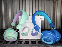 Pair of kids headphones side by side next to original packaging. One is teal with purple accents, the other is blue with teal accents. Both headphones are on-ear style with cushioned ear cups and adjustable headbands.