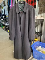 Photo of long dark men's coat hanging on a hanger, showing front button closure, collar, sleeves, and side pockets, inside a warehouse or storage setting.