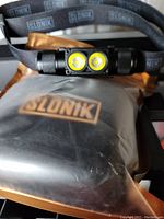 Photo showing the Slonik dual LED bulb headlamp attached to an adjustable black elastic headband, resting on top of a black carrying case with Slonik logo.