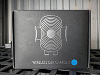 Black box packaging showing a white line drawing of the wireless car charger and stand with grip arms and circular center, labelled 'Wireless Car Charger' with a blue dot sticker.