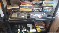 Photo showing stacked VHS tapes and some office supplies like tape rolls, pens and stapler, also photo paper pad, all on a black shelf.