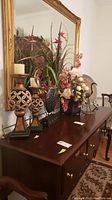 Wide shot showing candle holders, artificial floral arrangements in vases, metal bird figurine, and decorative plate on wooden furniture below large mirror.