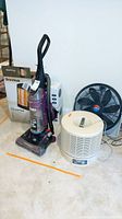 View of Eureka vacuum, OmniHeat and EcoHeat heaters in box, Lakewood Cool Operator fan against wall on carpet floor.