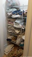 Wide shot of metal pantry shelves stacked with multiple folded linens including towels, pillows, drapes, and bath mats in a small closet space.