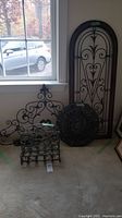 Three metal artworks near window: magazine holder, arched panel, and round medallion on floor